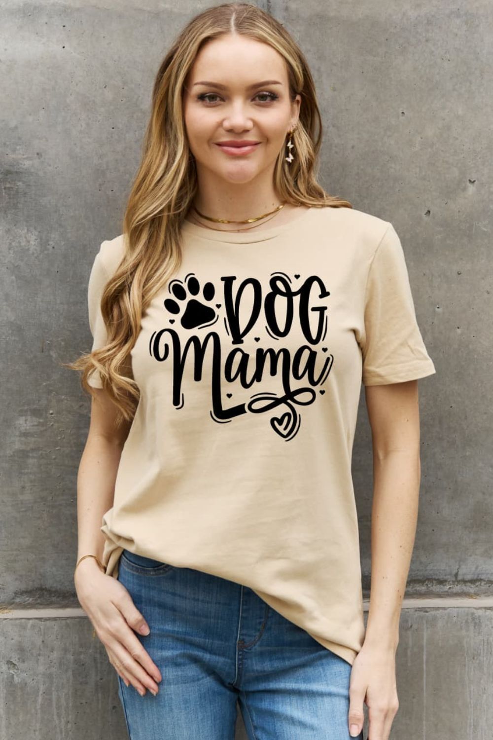 A woman in a beige "Dog Mama" t-shirt with a paw print and heart stands against a textured wall.