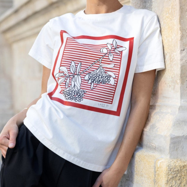A woman wearing a white t-shirt featuring a red-striped square frame with black and white floral line art, styled casually with black pants.