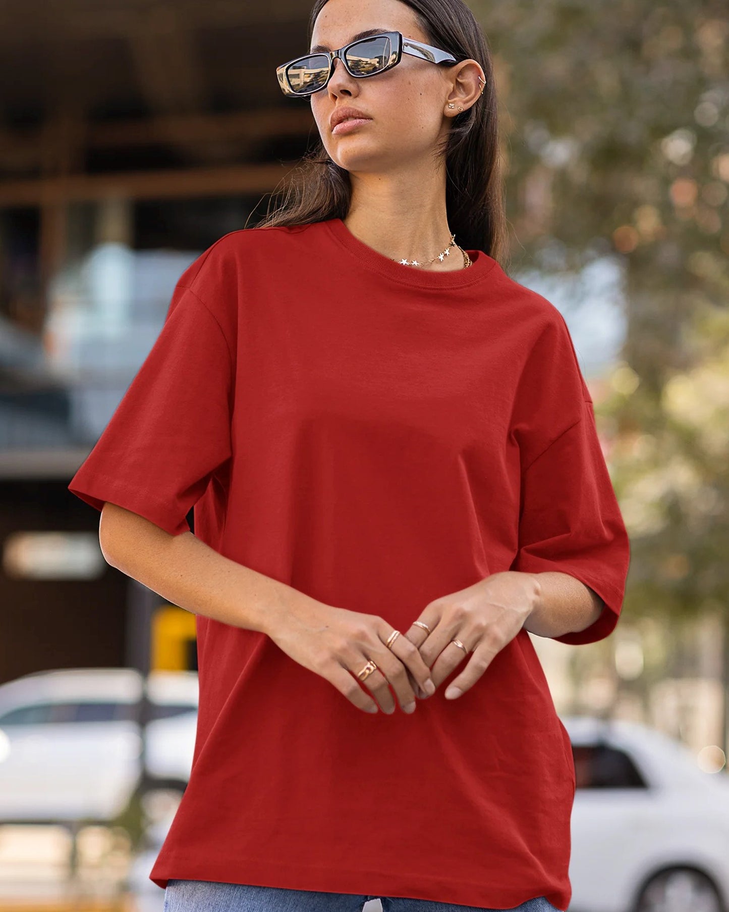 A person with dark hair and sunglasses wears a red oversized t-shirt, standing outdoors with hands at their waist.