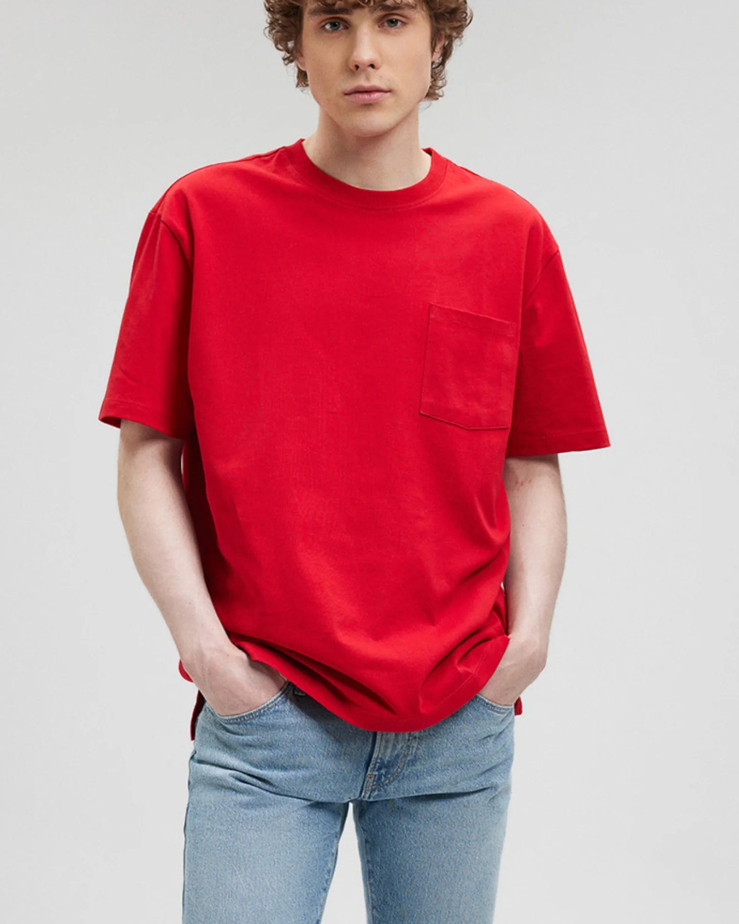 A person with light-colored hair wears a red t-shirt with a pocket and light wash jeans.