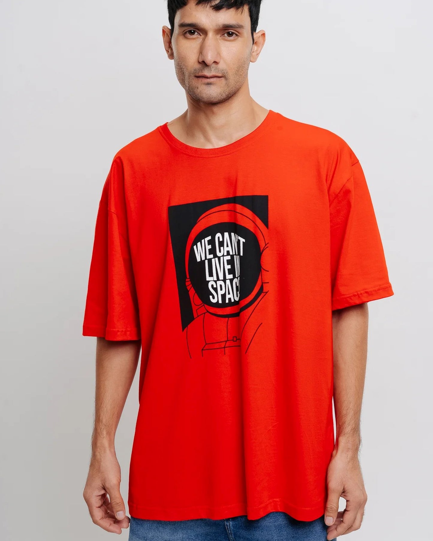 A man with dark hair wears a red t-shirt with a graphic and the text "We Can't Live In Space."