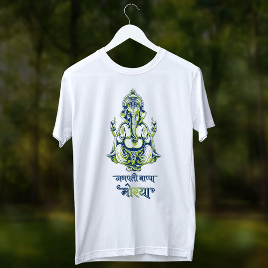 A white T-shirt featuring a detailed Lord Ganesha graphic in blue and green tones, along with Marathi text "गणपती बाप्पा मोरया." The shirt is displayed on a hanger against a blurred green outdoor background.
