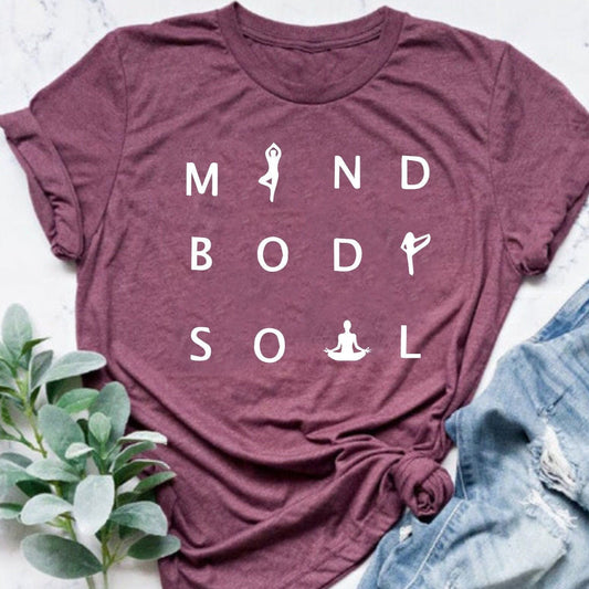 Maroon t-shirt with "MIND BODY SOUL" text and yoga pose symbols, styled with jeans and plant.