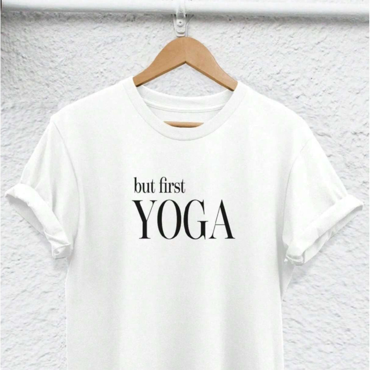 White t-shirt with "but first YOGA" text, hung on a hanger against a textured wall.