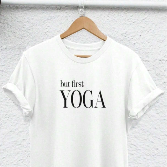 White t-shirt with "but first YOGA" text, hung on a hanger against a textured wall.