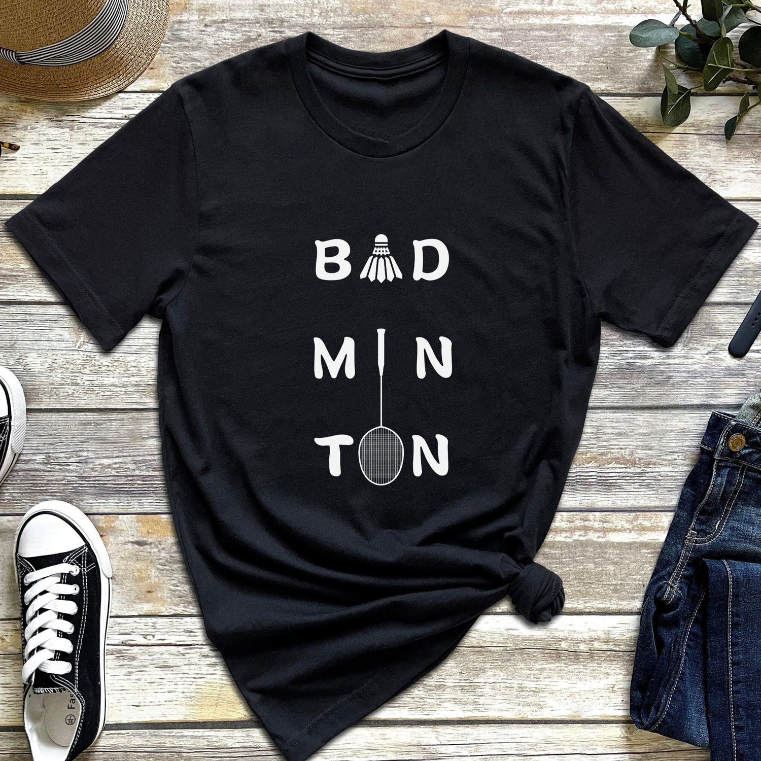 Black t-shirt with "Bad Min Ton" text and a simple shuttlecock graphic, styled with shoes, hat, and jeans.