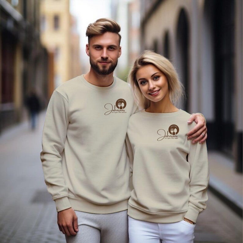 A man and woman stand close together wearing matching cream sweatshirts with a small logo on the chest.