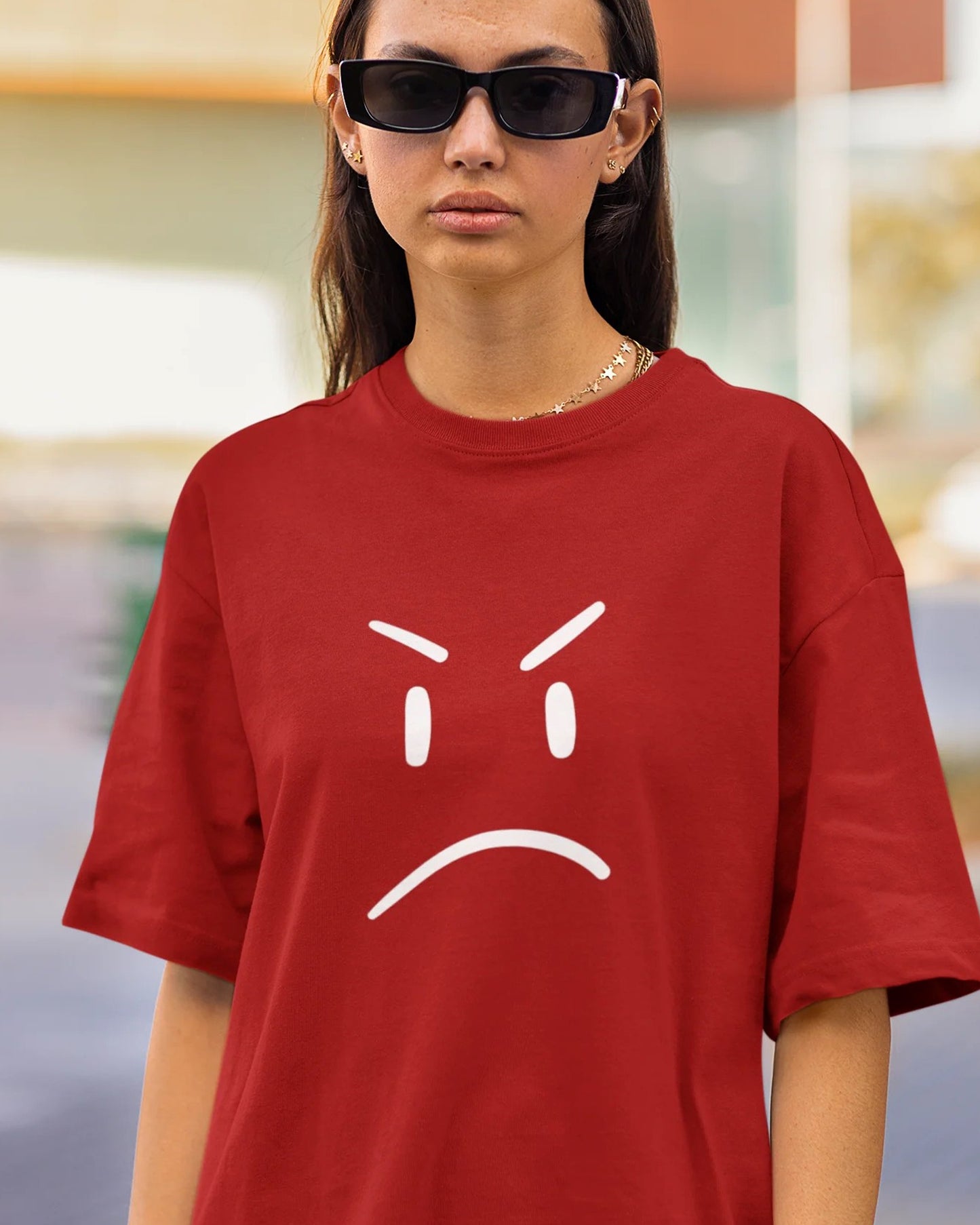 A young person with curly hair holds a black object to their ear, wearing a red t-shirt with a white angry face graphic.