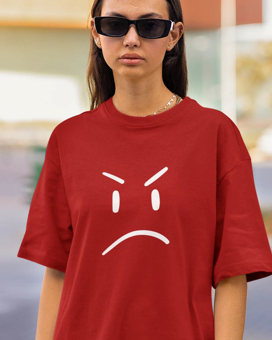 A young person with curly hair holds a black object to their ear, wearing a red t-shirt with a white angry face graphic.