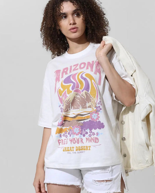 A person with curly brown hair wears a white t-shirt with a retro desert graphic and text, paired with white shorts.