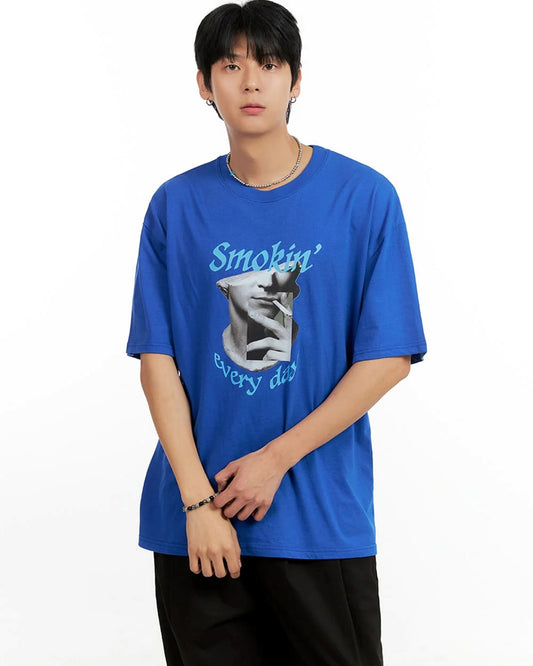 A young man with dark hair wears a blue t-shirt with a graphic print and text, paired with black pants.