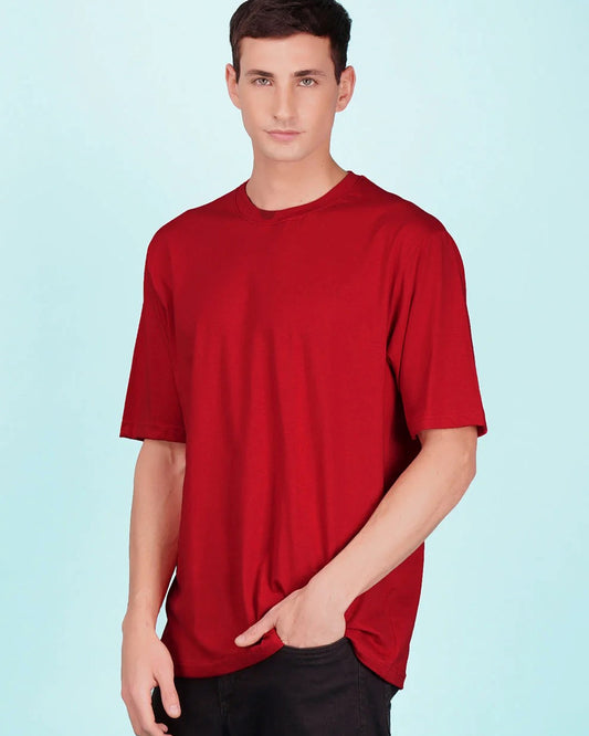 A man with dark hair and light eyes wears a red t-shirt and dark pants against a light blue background.