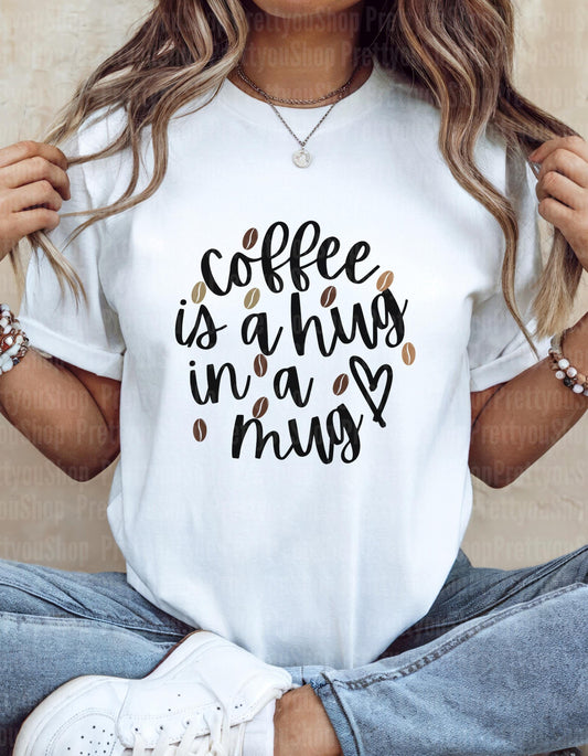 White t-shirt with "Coffee is a hug in a mug" text and coffee bean graphics. A comforting design for coffee enthusiasts.