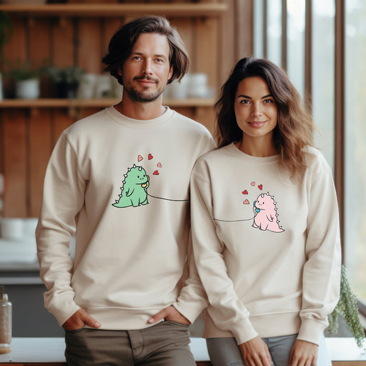 A man and a woman wearing matching beige sweatshirts with cartoon dinosaurs and hearts, posing indoors.
