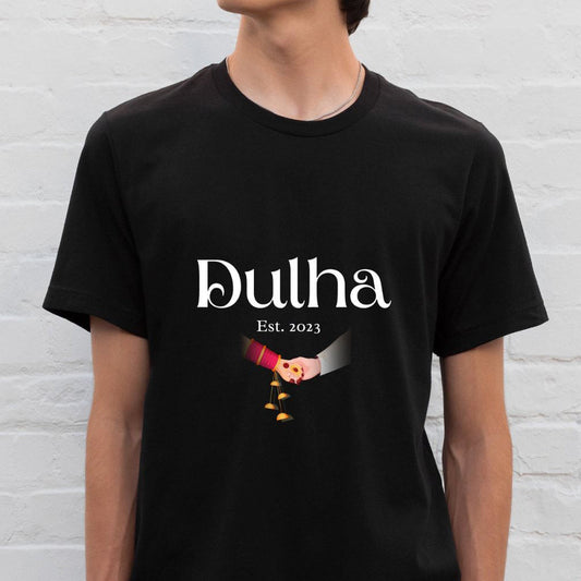 Black cotton t-shirt with "Dulha" in white script, "Est. 2023" below, and a graphic of hands exchanging gold coins, likely indicating a wedding ritual.