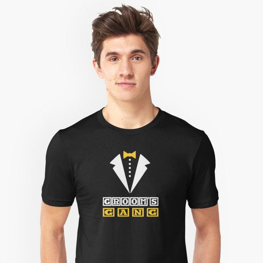 Black cotton t-shirt featuring a white tuxedo graphic with a gold bow tie and buttons, above yellow and white block letters spelling "GROOMS GANG".