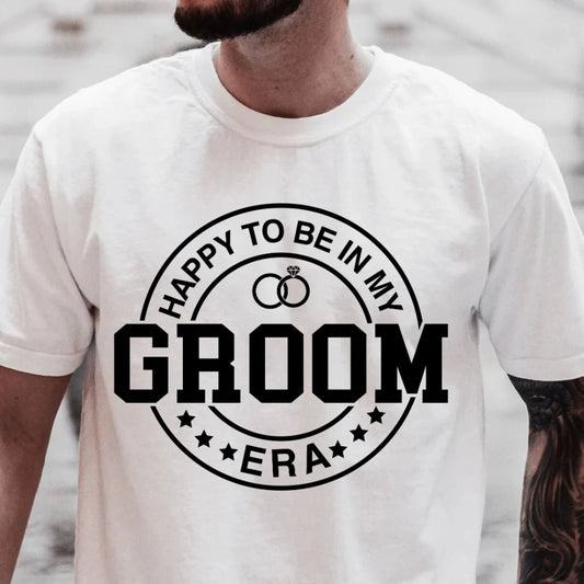 White cotton t-shirt with a black circular design featuring wedding rings, "HAPPY TO BE IN MY" at the top, "GROOM" in bold, and "ERA" at the bottom.