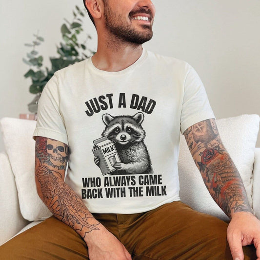 A man wearing a cream-colored t-shirt with a raccoon holding a milk carton and the text "JUST A DAD WHO ALWAYS CAME BACK WITH THE MILK".