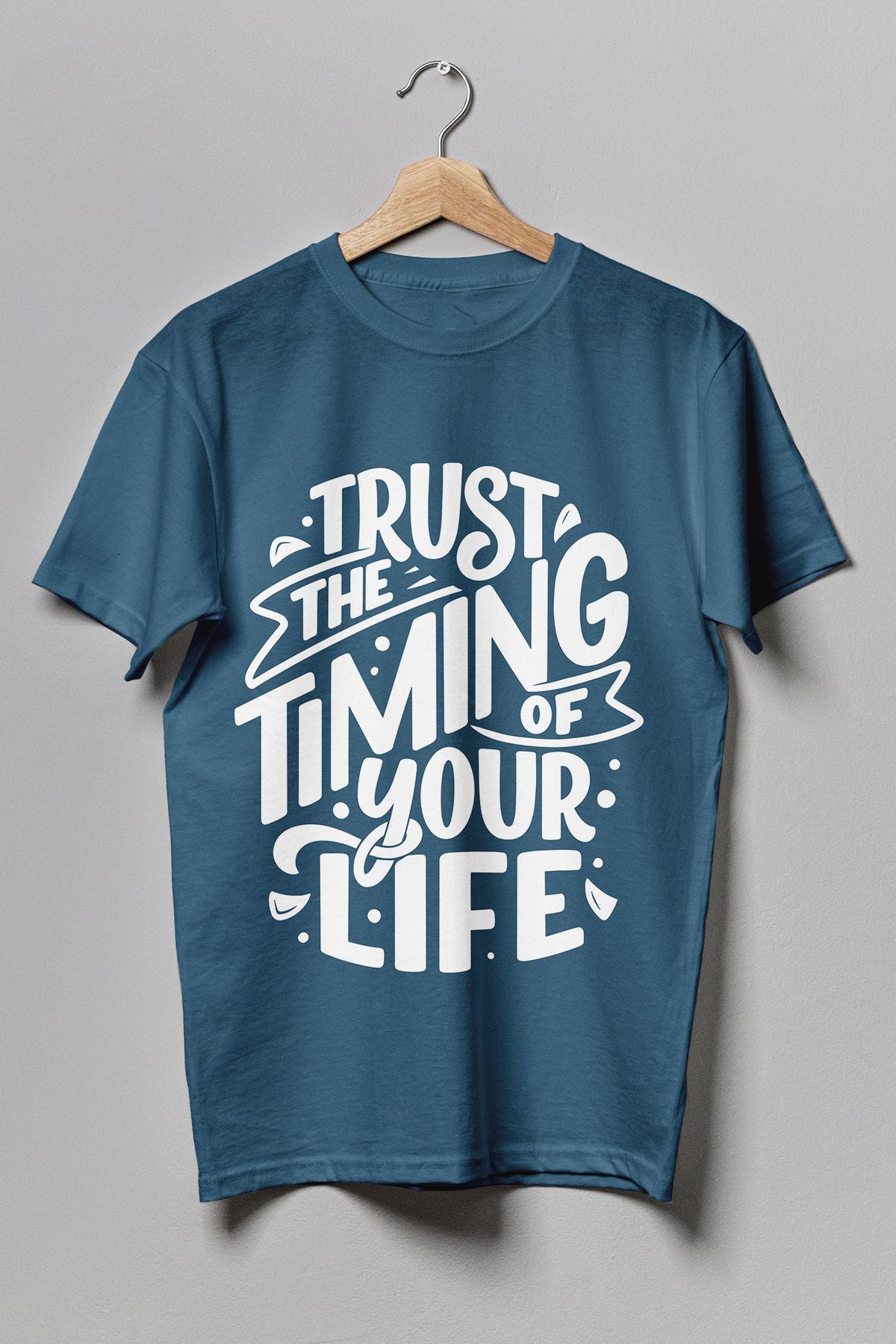 Teal t-shirt hanging on a wire hanger against a gray background, featuring white stylized text "TRUST THE TIMING OF YOUR LIFE".