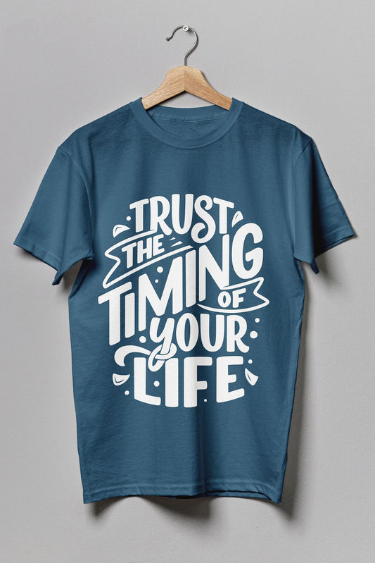Teal t-shirt hanging on a wire hanger against a gray background, featuring white stylized text "TRUST THE TIMING OF YOUR LIFE".