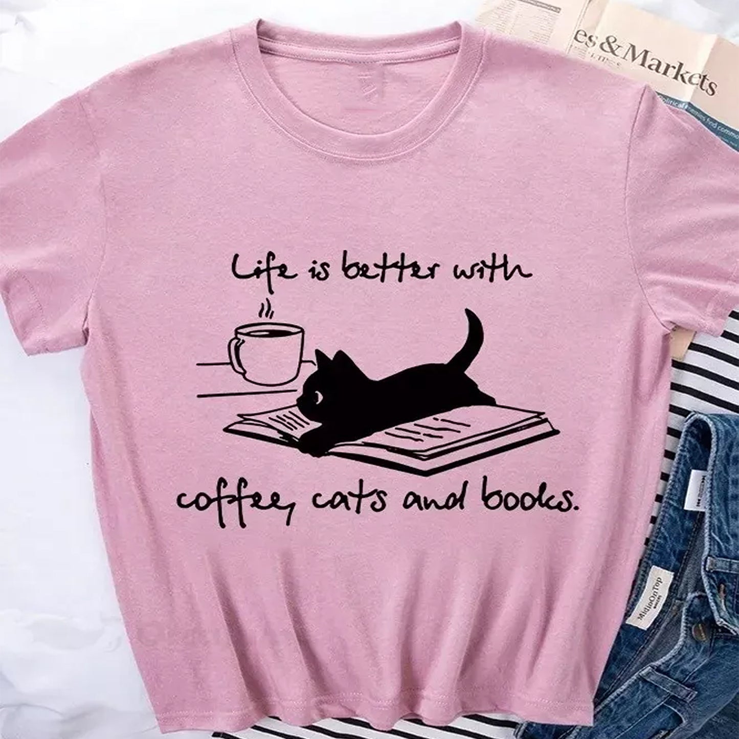 Pink t-shirt with a graphic of a cat on a book, coffee cup, and "Life is better with coffee, cats and books" text.  A cozy design for book lovers.