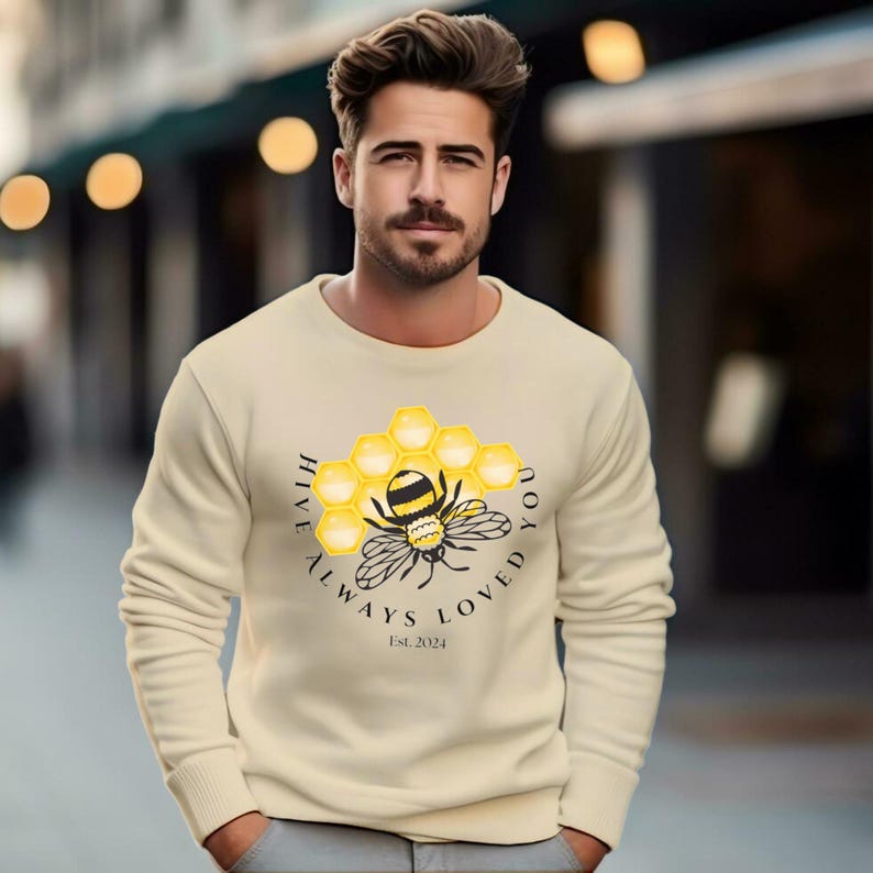 A smiling man and woman stand together wearing matching cream sweatshirts featuring a bee, honeycomb, and text.