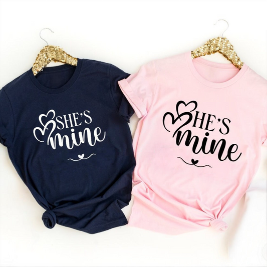 A navy blue t-shirt reading "She's Mine" with a heart and a light pink t-shirt reading "He's Mine" with a heart.