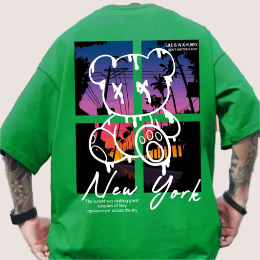 A green oversized t-shirt with a graphic of a teddy bear, "New York" text, a sunset scene, and the phrase "Life is in a hurry, don't miss the sunset."