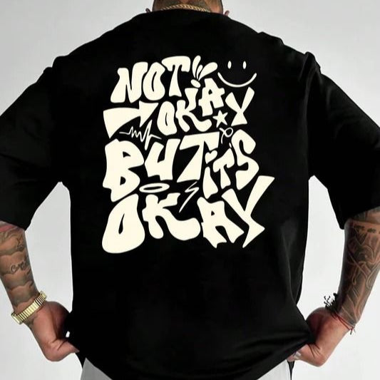 Black oversize t-shirt with the phrase "NOT OKAY BUT IT'S OKAY" in a stylized, graffiti-inspired white font on the back. The graphic also includes a smiley face and other small doodle-like elements. 