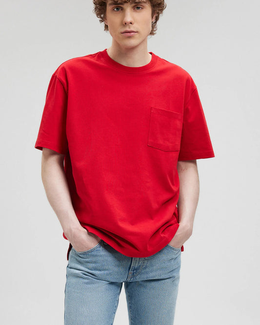  A person with light-colored hair wears a red t-shirt with a pocket and light wash jeans.