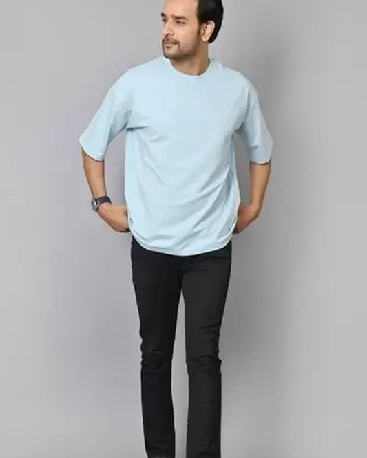 A man wearing a light blue oversized t-shirt and black pants stands with hands in pockets.
