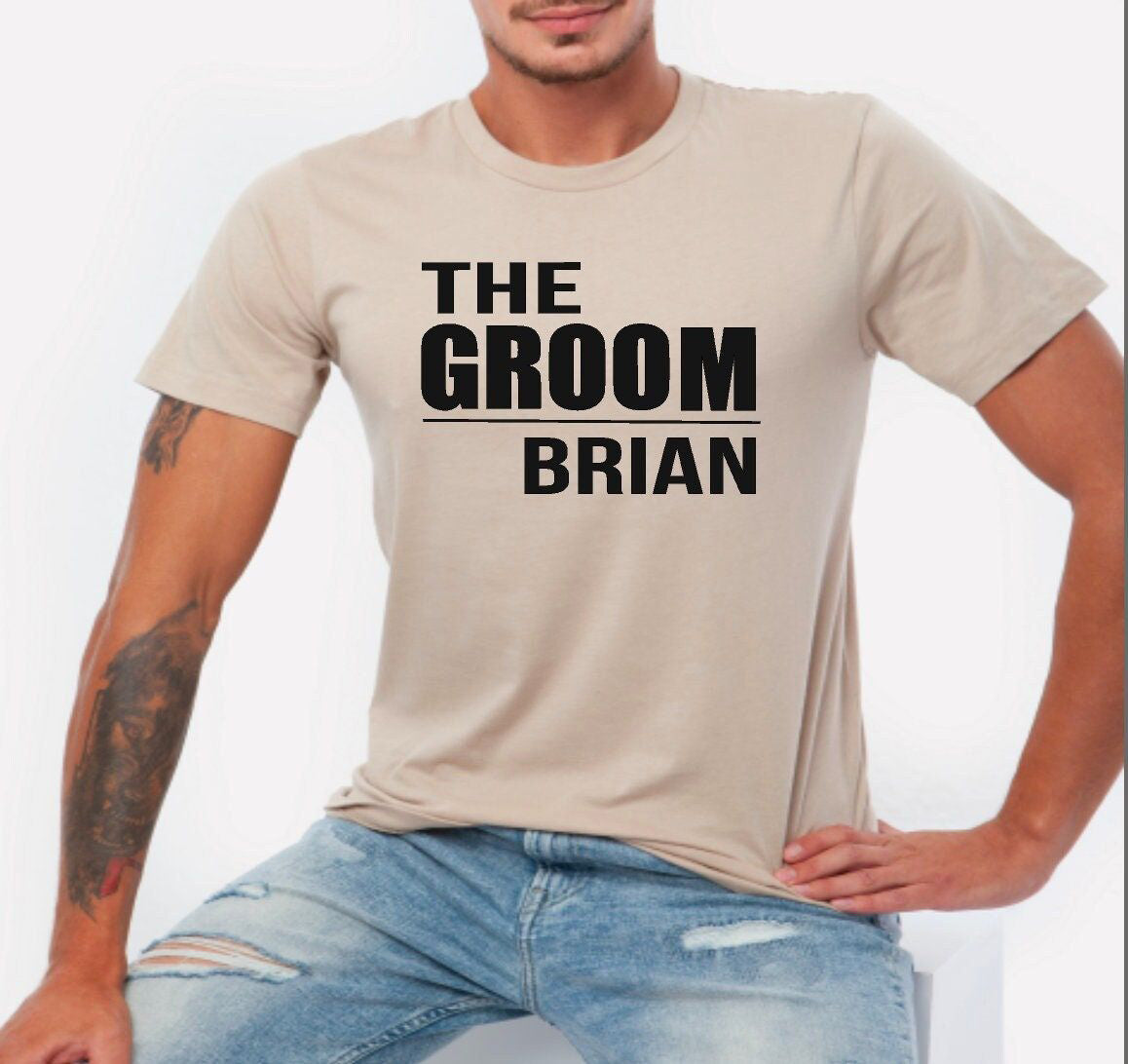 Light brown cotton t-shirt with black printed text "THE GROOM" above [Name] in bold, worn by a person with a tattoo.