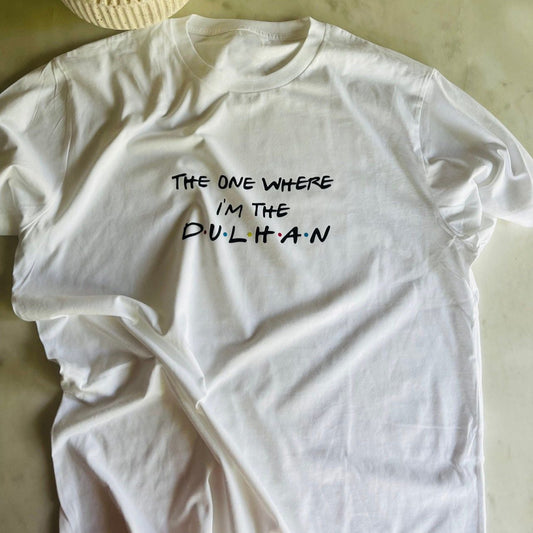 White cotton t-shirt with black text "THE ONE WHERE I'M THE DULHAN", likely a bride's celebratory t-shirt. The word "Dulhan" suggests a South Asian cultural context for the wedding.