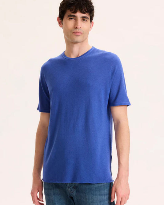 A man with dark hair wears a bright blue t-shirt and jeans against a white background.