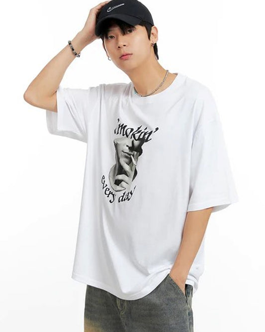 A young person with dark hair wears a white t-shirt with a graphic print and text, paired with gray pants.