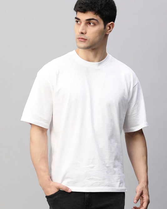  A man with dark hair wears a plain white oversized t-shirt and dark pants against a light gray background.