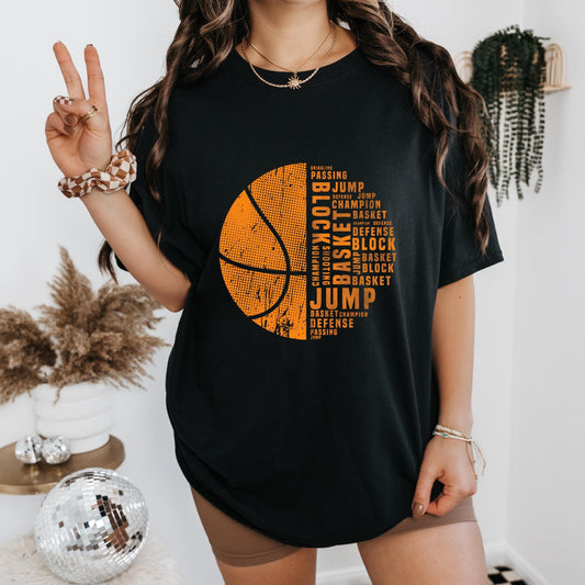 Black oversized T-shirt featuring an orange half-basketball design filled with basketball-related words like 'Jump,' 'Block,' 'Defense,' and 'Champion,' worn by a woman posing with a peace sign in a stylish indoor setting with a disco ball and modern decor.